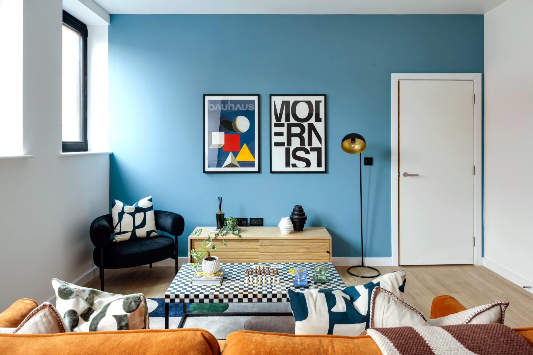 Blue feature wall luxury living room with Bauhaus posters, a checkered coffee table, and burnt orange velvet sofa.