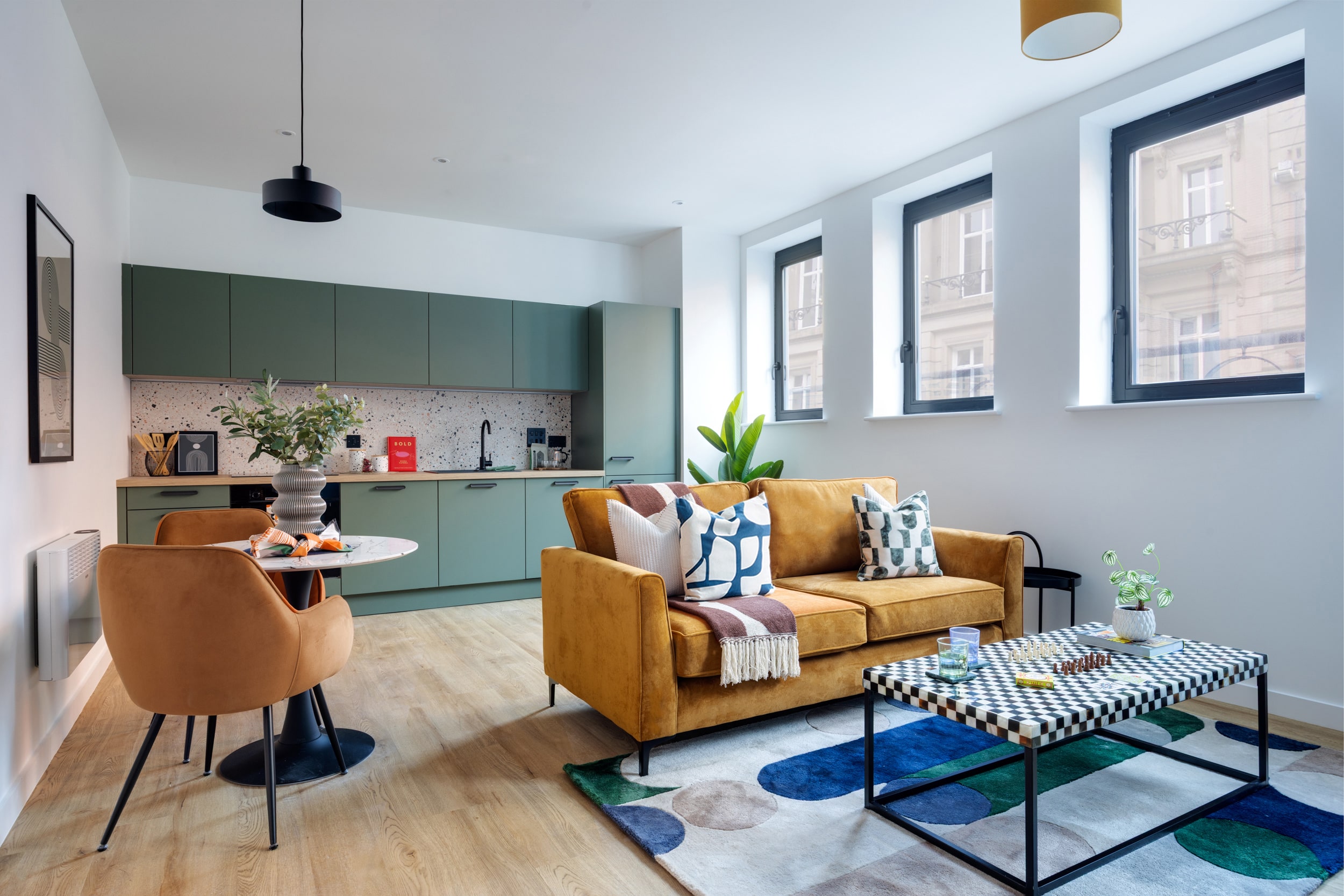 Open-plan apartment interior featuring a sage green modern kitchen with terrazzo splashback, an ochre velvet sofa, and a geometric checkered coffee table.