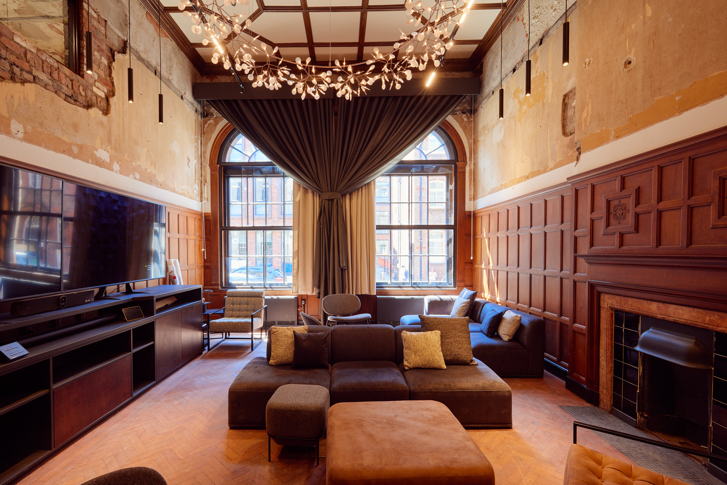 A luxury executive lounge at Duke & Parr, Liverpool, featuring original dark wood wall paneling, a historic fireplace, coffered ceilings, and a large-scale AV media unit, balanced with raw exposed plaster walls and designer lighting.