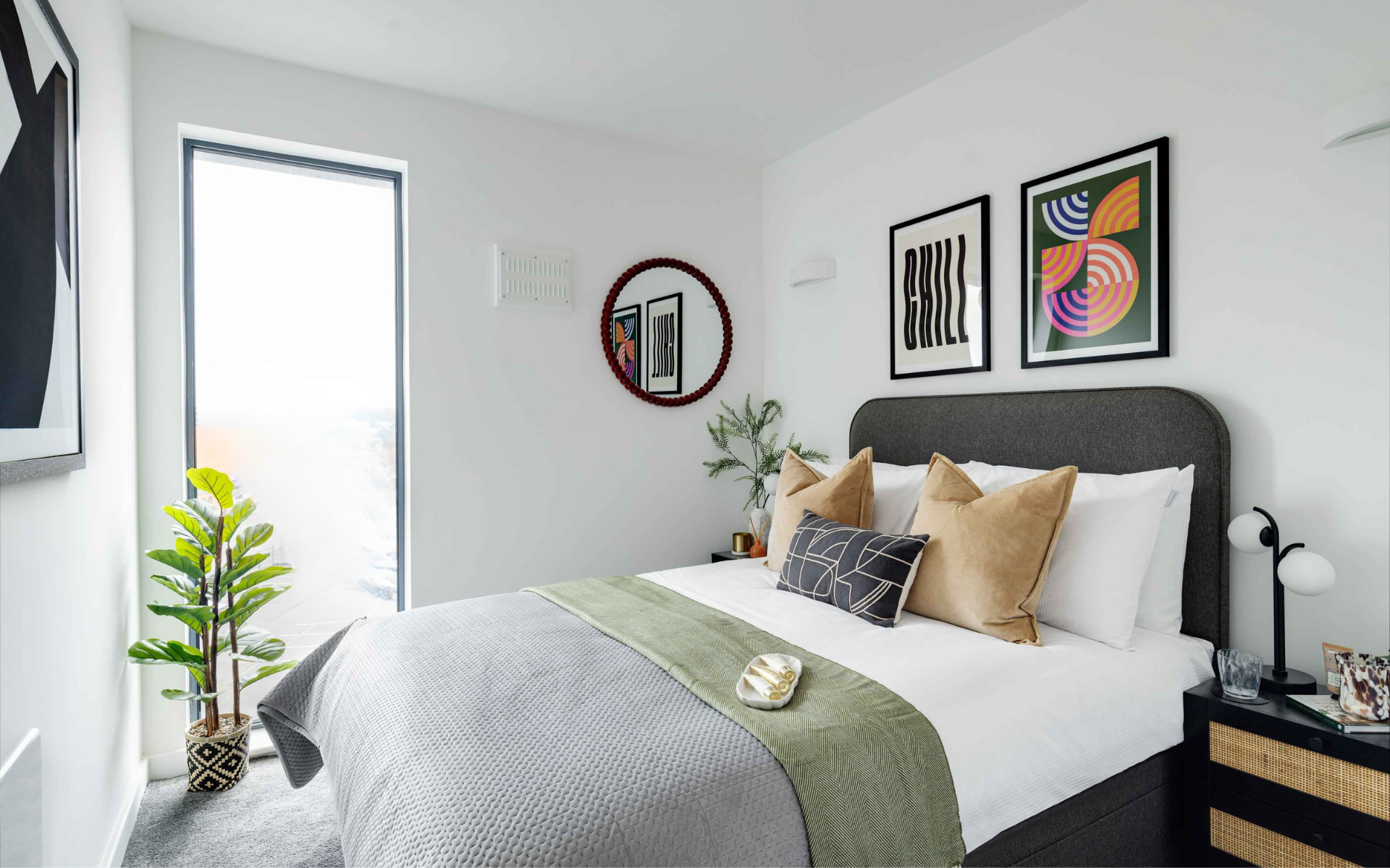 Modern apartment bedroom interior styled by CERT Property, featuring a double bed with a dark grey upholstered headboard, beige cushions, a green bed throw, contemporary framed art prints, a circular wall mirror, and a rattan bedside table.