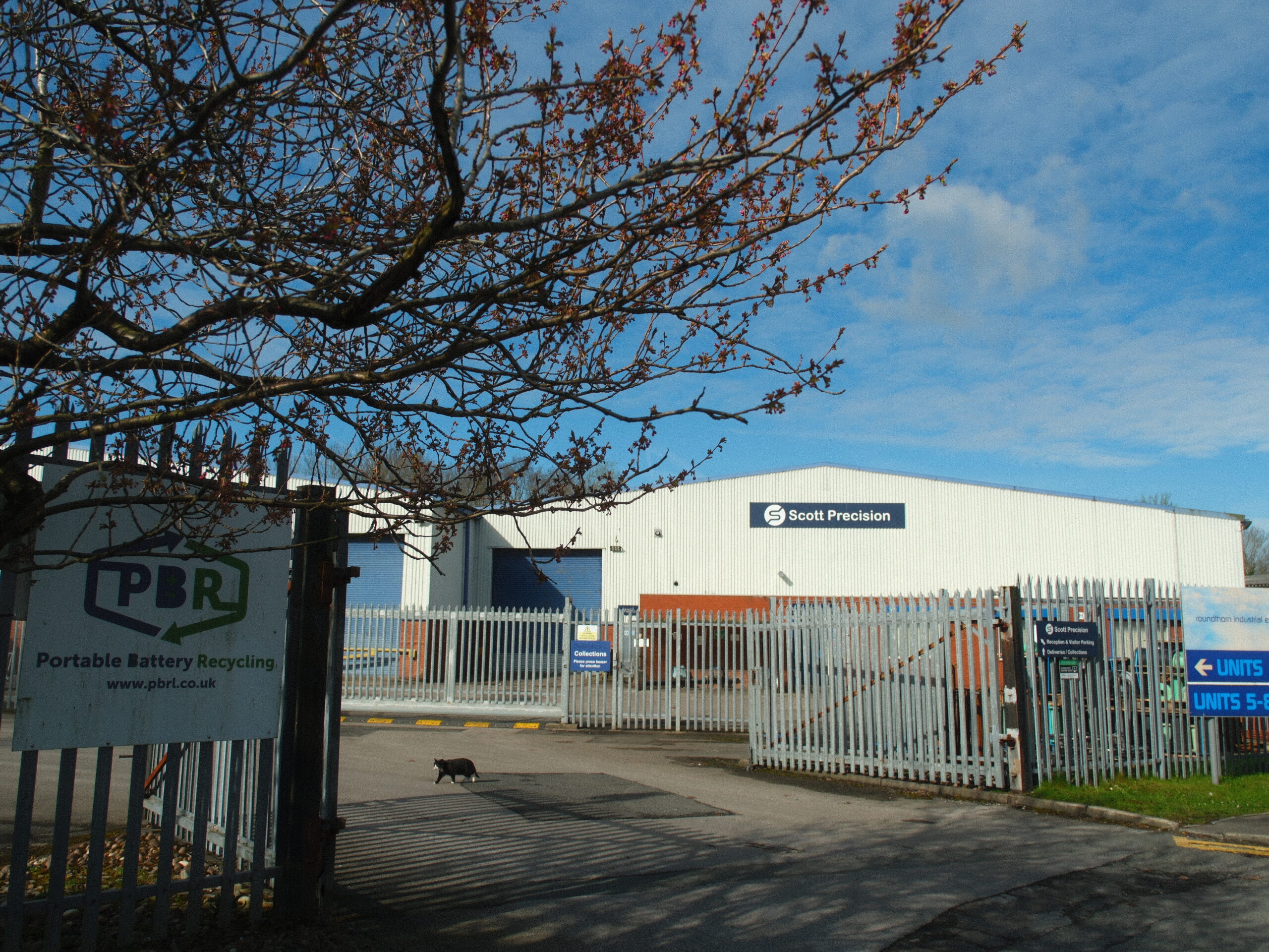 External view of the gated entrance to Roundthorn Industrial Estate in Wythenshawe, showing a PBR Portable Battery Recycling sign on the fence and industrial warehouses under a blue sky.
