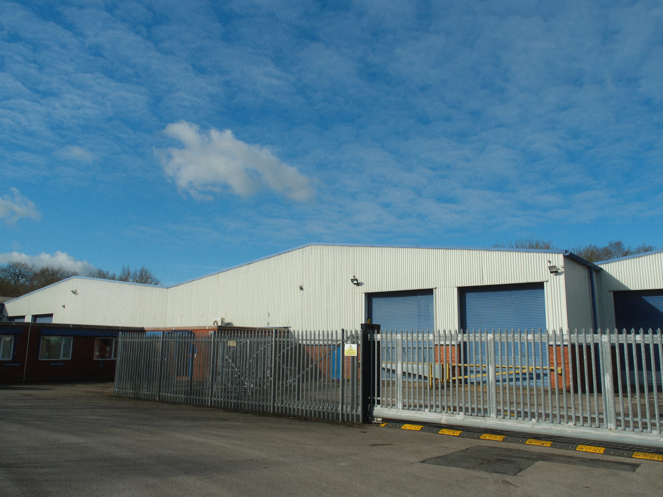 Angled external view of commercial warehouse units at Roundthorn Industrial Estate, featuring white steel cladding, blue roller shutter loading bays, and a secure metal perimeter fence.