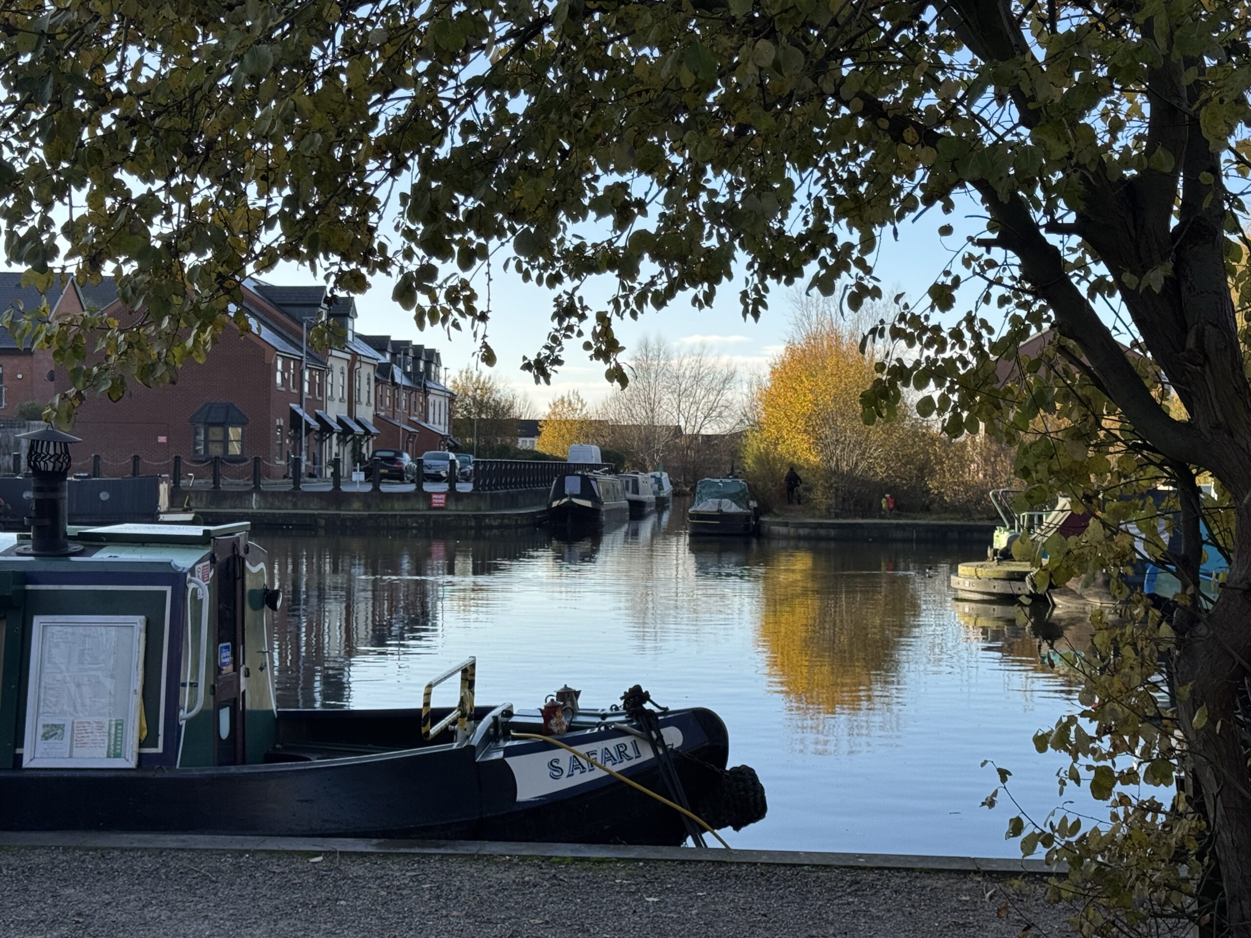 A scenic, tree-framed view of the Ashton Canal basin in Droylsden, showing traditional narrowboats moored along the banks and autumn trees reflecting in the still water.