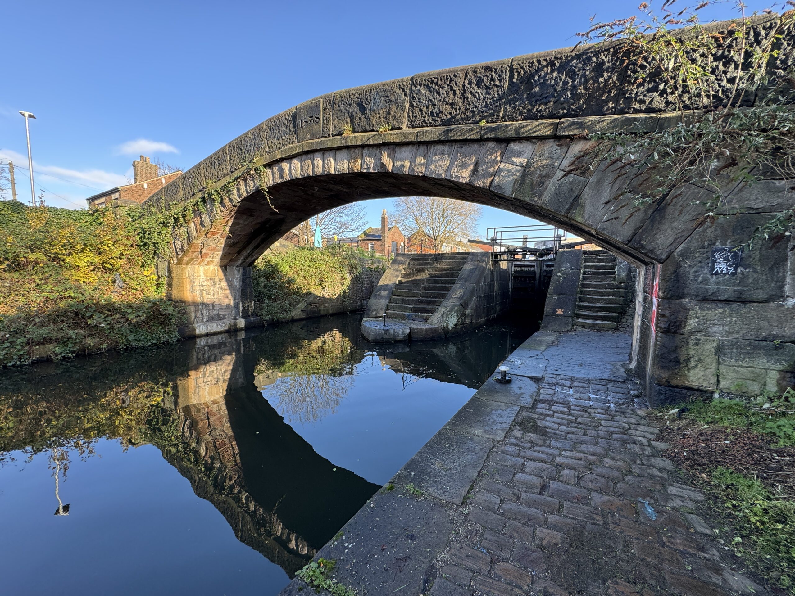 A scenic daytime view of a historic arched stone canal bridge over quiet water, revealing a lock gate system with stone steps reflecting in the canal pool.